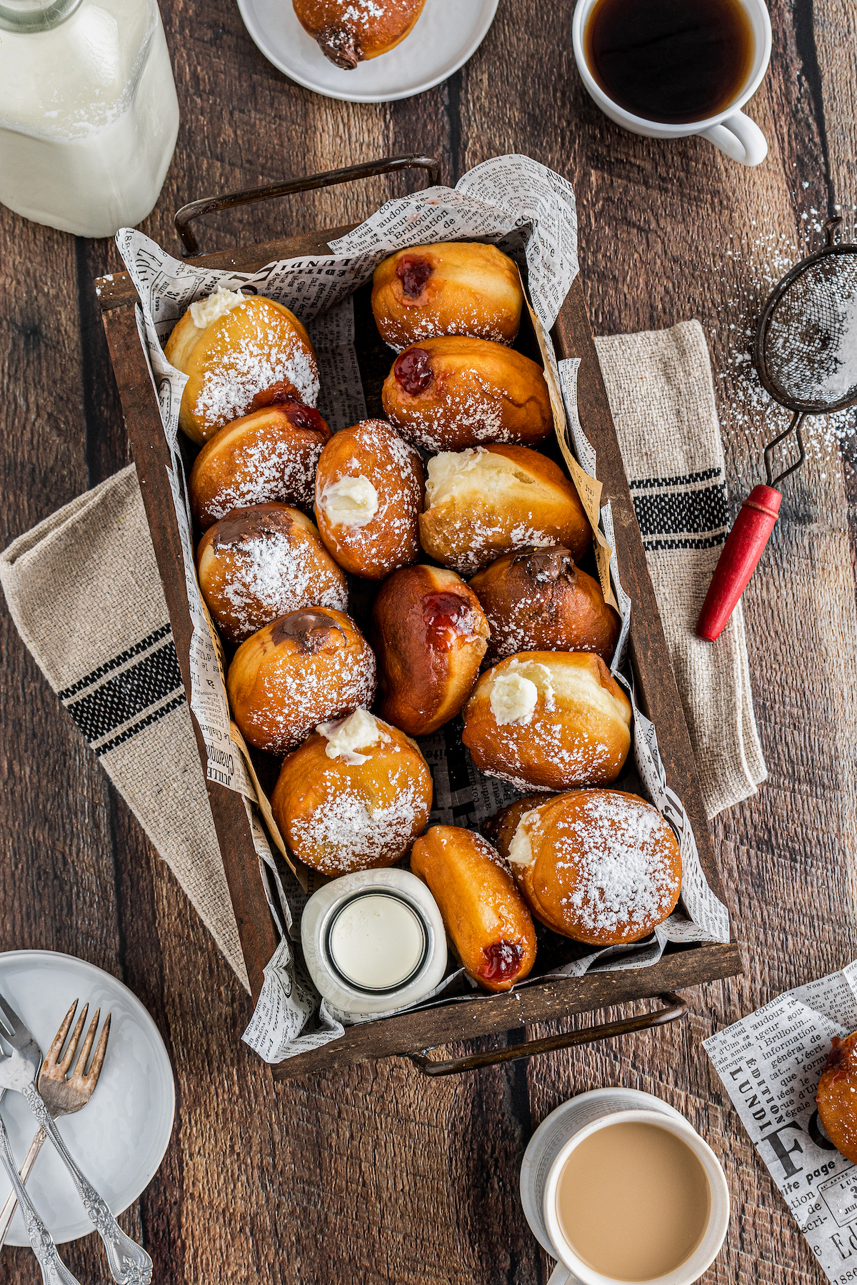 A rustic tray full of donuts, set on a wooden table, surrounded by coffee cups, saucers, napkins, powdered sugar, and a sieve.
