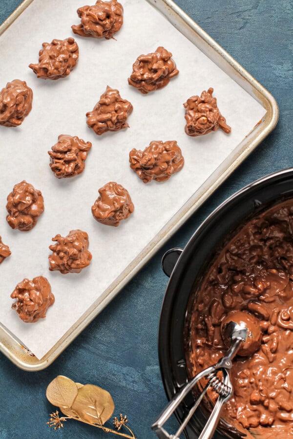 Simply toss all of the ingredients into the crockpot, stir a couple of times, scoop, and bam! Homemade Peanut Butter Pretzel Crockpot Candy! Crockpot candy in a crockpot being scooped out with a cookie scoop onto parchment paper on a baking sheet.