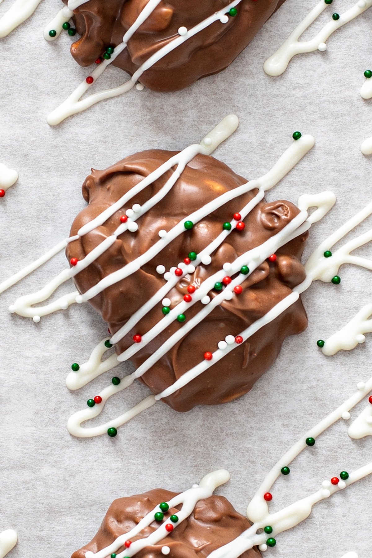 Simply toss all of the ingredients into the crockpot, stir a couple of times, scoop, and bam! Homemade Peanut Butter Pretzel Crockpot Candy! Crockpot candy on white parchment paper being drizzled with white chocolate and topped with Christmas sprinkles.
