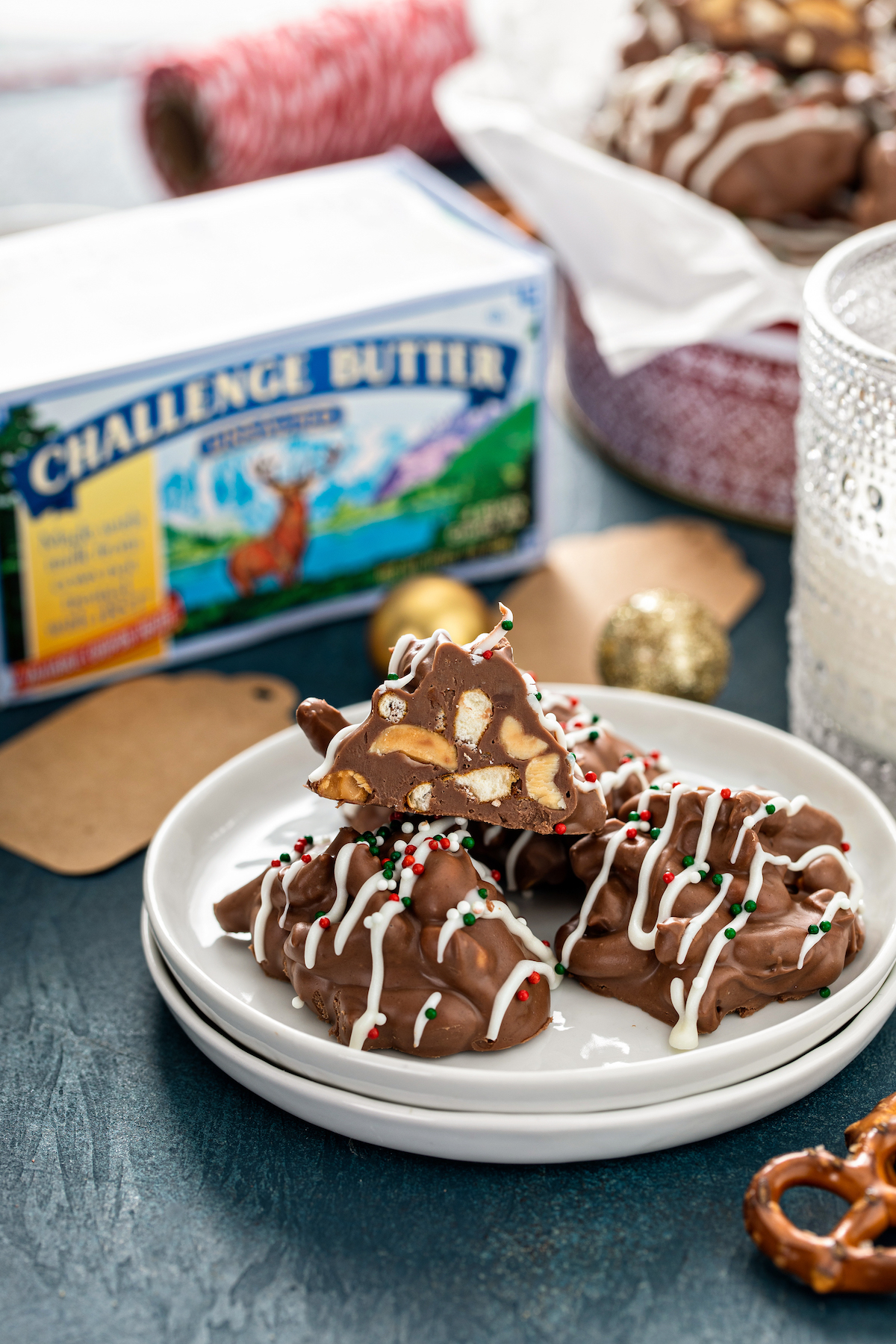 Candy clusters stacked on a white plate with pretzels, milk and a box of butter in the background.