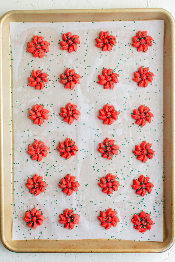 Unbaked red spritz cookies with green sprinkles on a cookie sheet.
