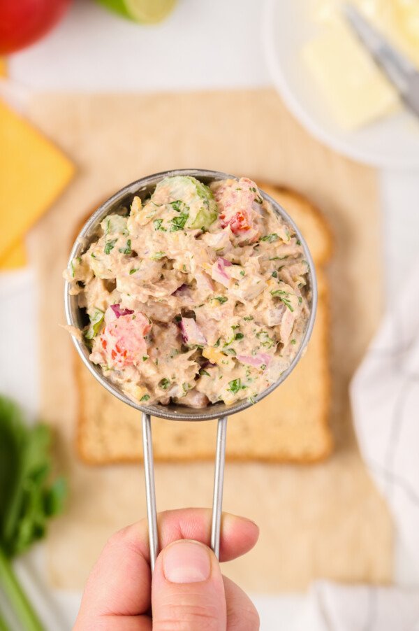 A stainless steel measuring cup full of tuna salad, with bread and other ingredients in the background.