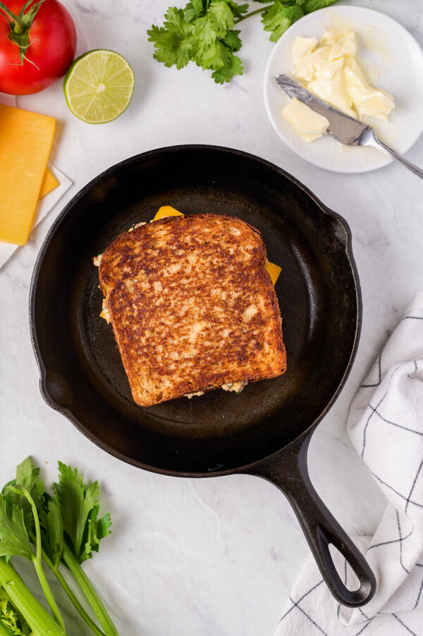 A grilled sandwich in a cast-iron pan. Tomato, cheese, butter, herbs, and other ingredients are arranged around the work surface.