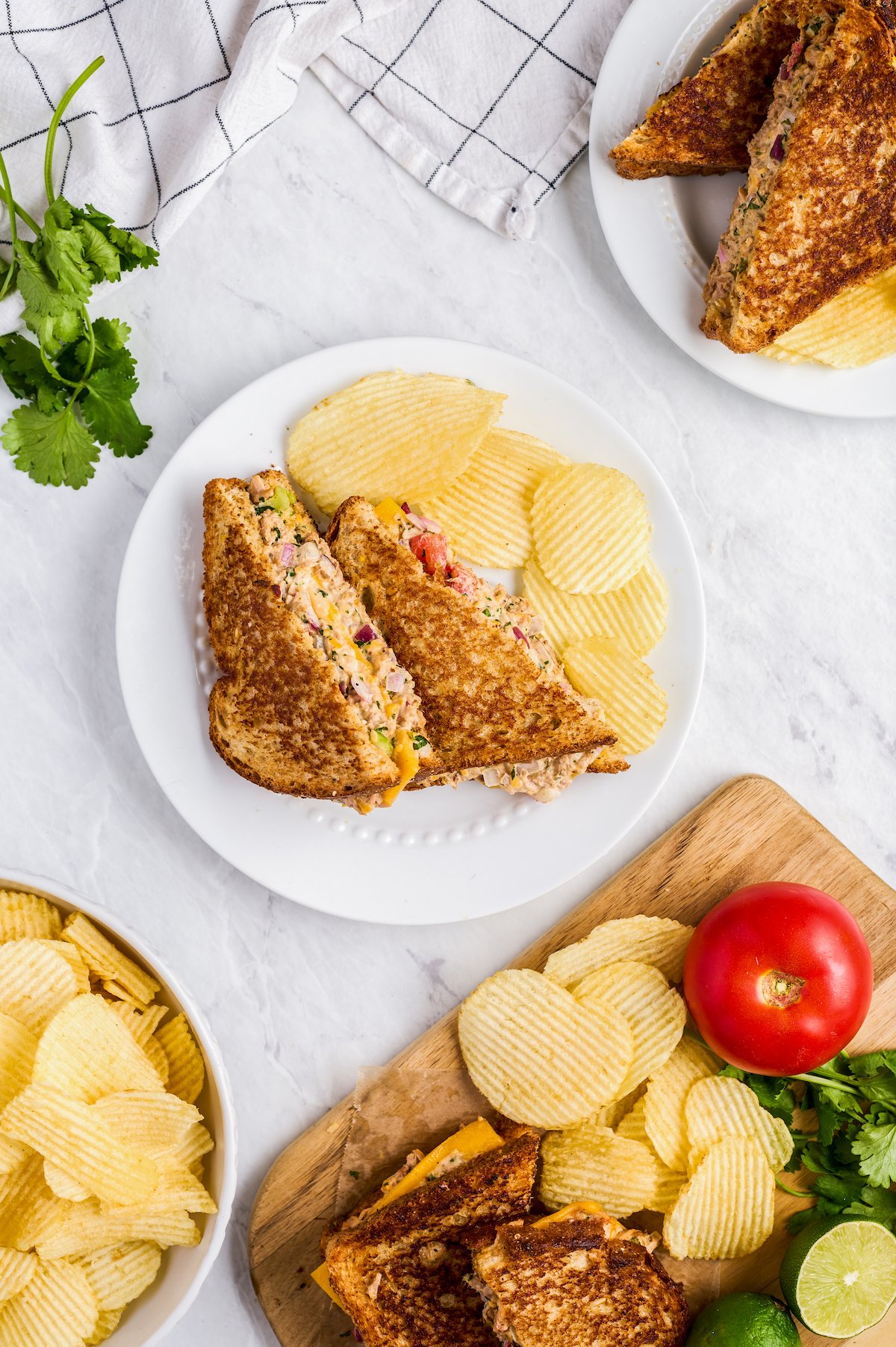 A tuna salad sandwich with chips on a white plate. Tomatoes, herbs, and other ingredients are arranged on the table as well.