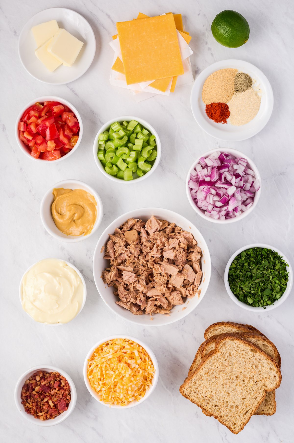 Left to right, from top: pats of butter, sliced cheese, lime, tomato, celery, garlic powder, onion powder, cayenne pepper, black pepper, mustard, red onion, mayonnaise, tuna, cilantro, bacon, cheese, bread.