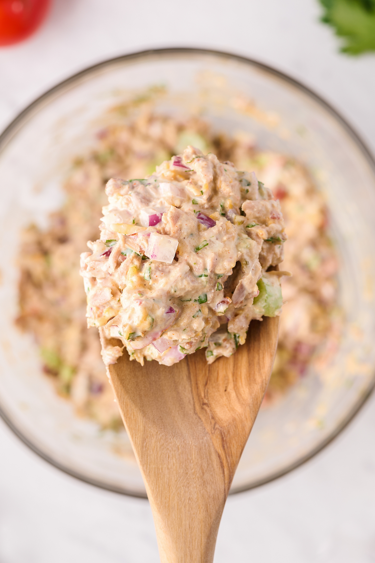 An overhead shot of tuna salad being lifted out of a glass bowl with a wooden spoon.