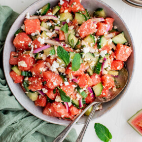 A white serving dish filled with watermelon feta salad. Two serving utensils are in the bowl, and a green napkin is on the table.