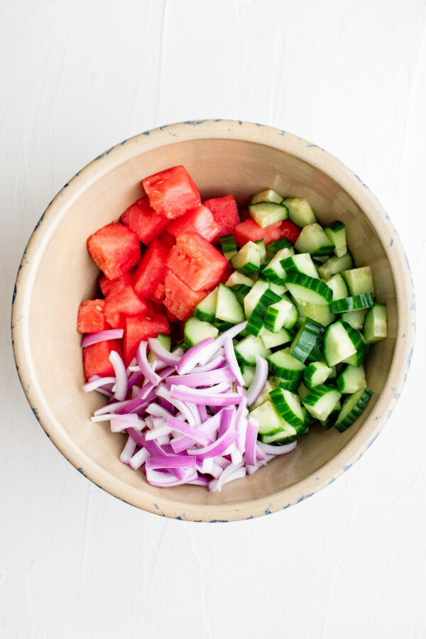 A large salad bowl with sliced red onion, cucumber, and watermelon cubes added.