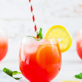 A small glass of watermelon lemonade, garanished with lemon slices, mint leaves, and a red-and-white striped straw.