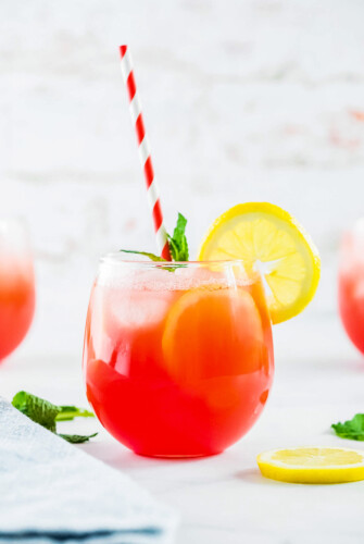 A small glass of watermelon lemonade, garanished with lemon slices, mint leaves, and a red-and-white striped straw.