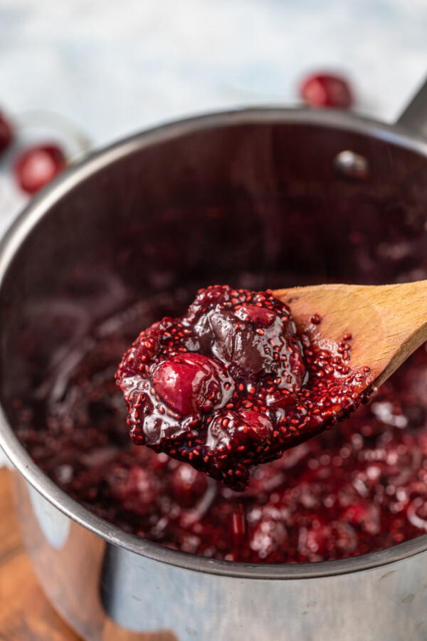 Cherry chia jam on a wooden spoon in a pot.