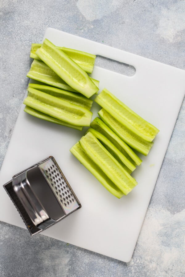 A cutting board with pieces of peeled, seeded cucumber and a box grater.
