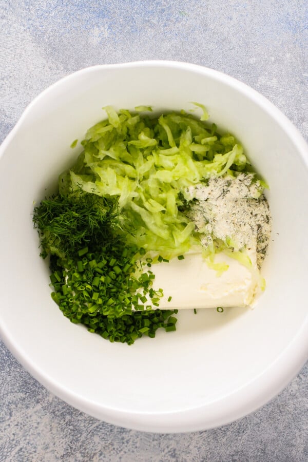 A mixing bowl filled with grated cucumber, ranch seasoning, cream cheese, and fresh herbs.
