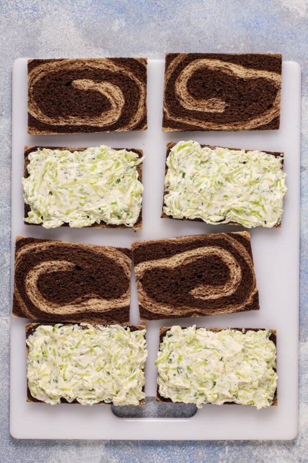 Slices of marble rye laid on a cutting board. Half of the slices are spread with creamy cucumber mixture.