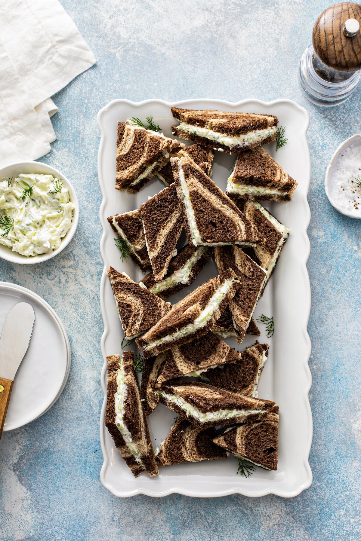 A rectangular white platter with triangular finger sandwiches on marble rye, next to a small bowl of cream cheese filling and a cheese knife.