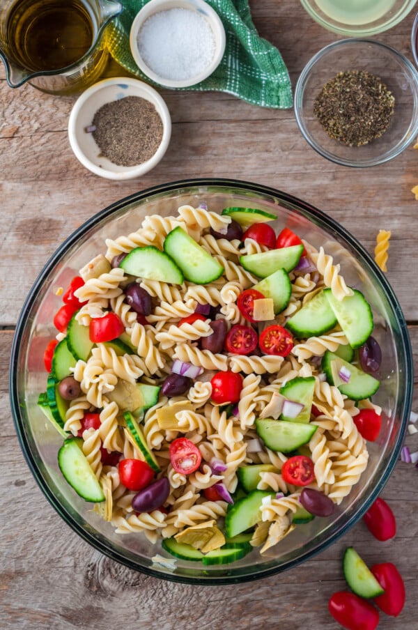 A bowl filled with pasta, red onion, tomatoes, olives, cucumbers and artichoke hearts.