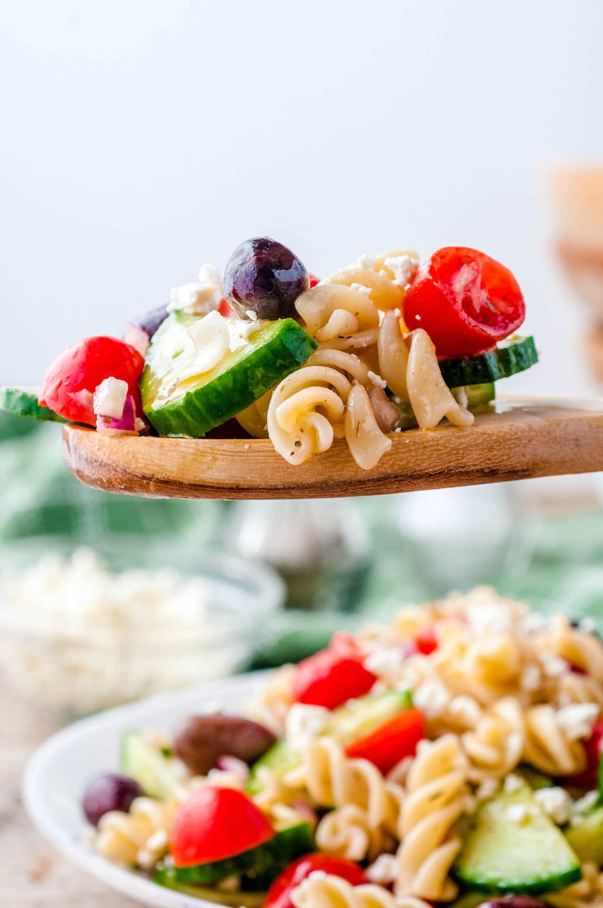 A wooden spoon scooping up a serving of greek pasta salad from a bowl.