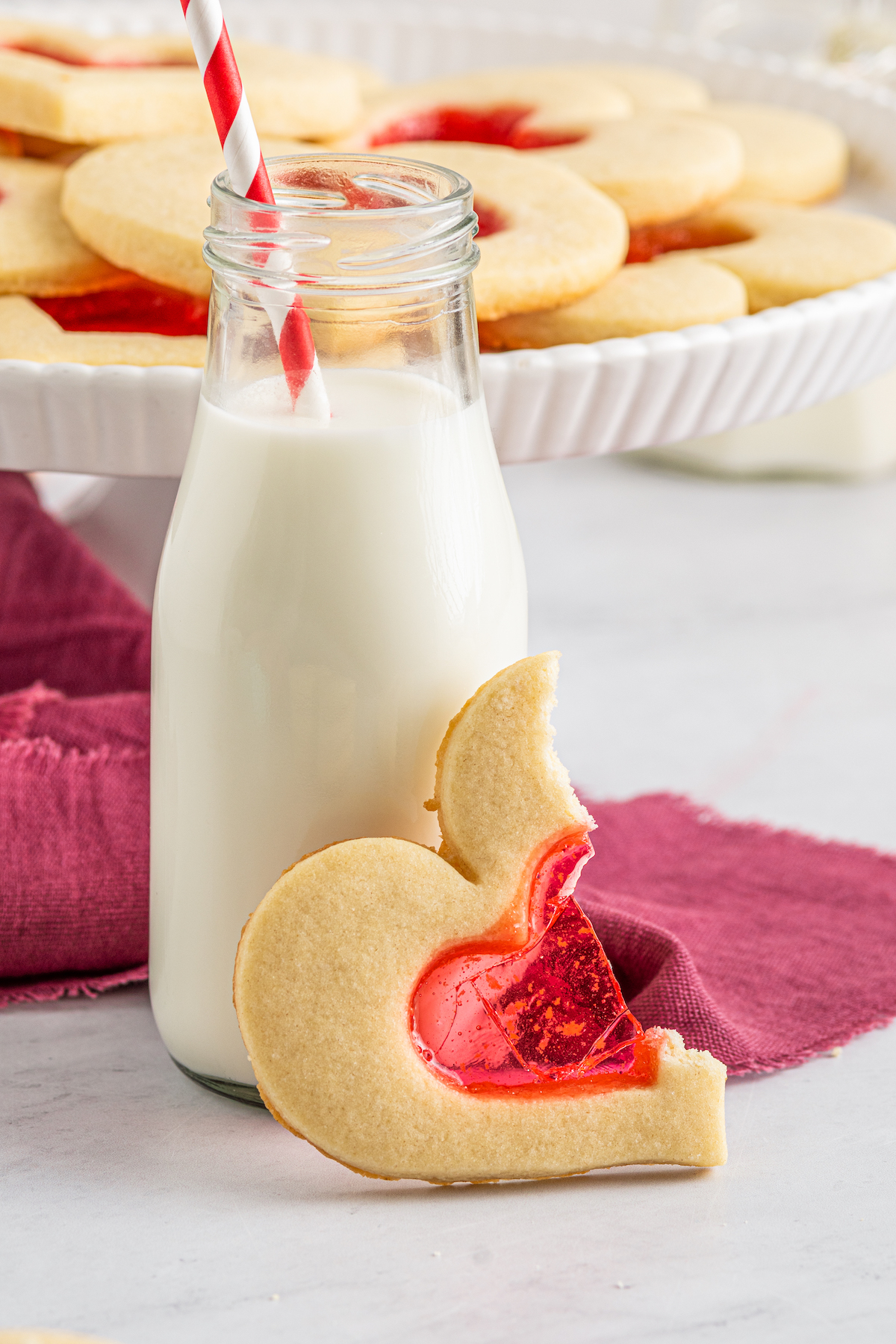 A heart-shaped cookie leaning against a bottle of milk with a straw. The cookie has been broken in half to show texture.