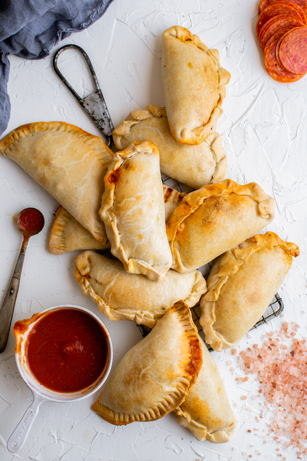 Eight empanadas piled on a wire rack, with a measuring spoon, a cup of pizza sauce, and a few pepperoni nearby.