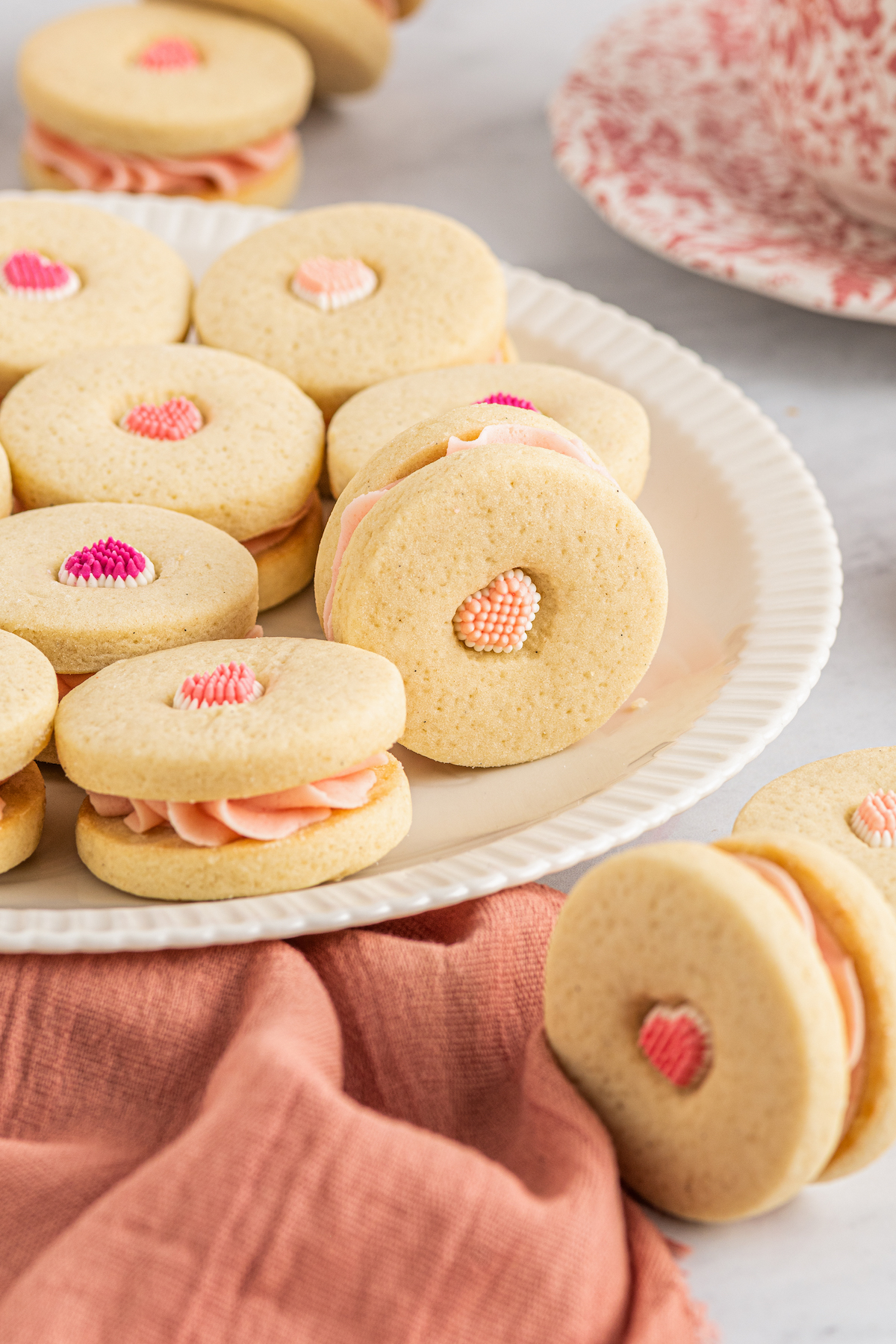 A white plate with vanilla sandwich cookies arranged on it in a single layer. A pink napkin and cookie are in the foreground of the shot.