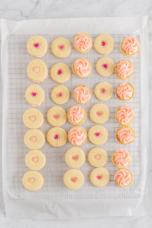 A cookie rack with cookies on it. Some are decorated with candy hearts, while others are topped with piped pink buttercream.