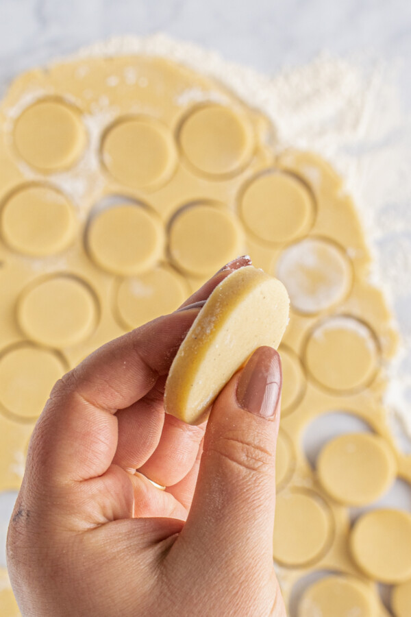 A woman's hand is holding a circle of unbaked cookie dough. In the background of the shot is a work surface with a flat sheet of dough. Cookies have been cut out of the dough.