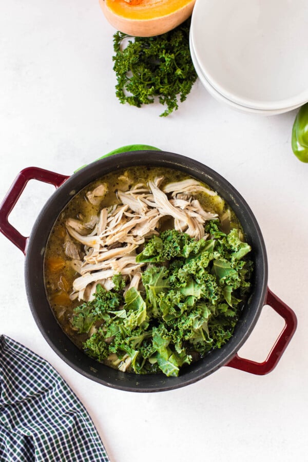 Chicken breasts shredded in broth with fresh kale added to the pot.