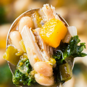 A spoon holding a bite of chicken and vegetable soup with kale and butternut squash.