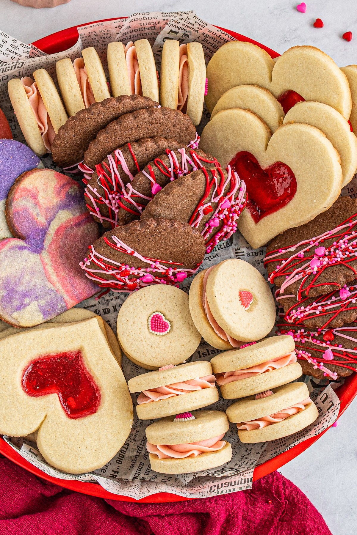 Heart-shaped cookies, sandwich cookies, marbled cookies, and chocolate iced cookies arranged on a red plate.
