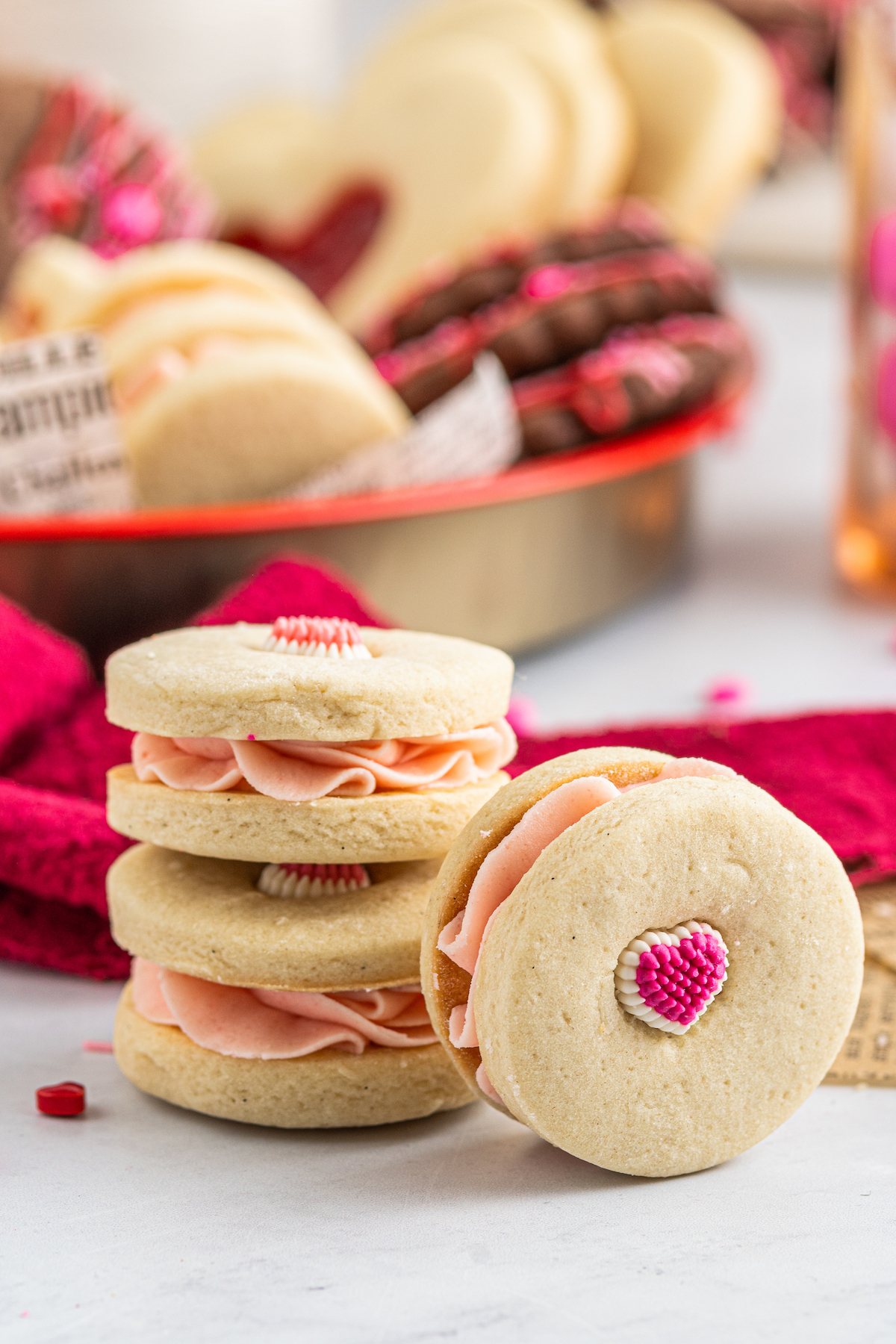 A platter of cookies is in the background of the shot. In the foreground are three sandwich cookies with piped icing in the centers and candy hearts pressed into the cookie dough.