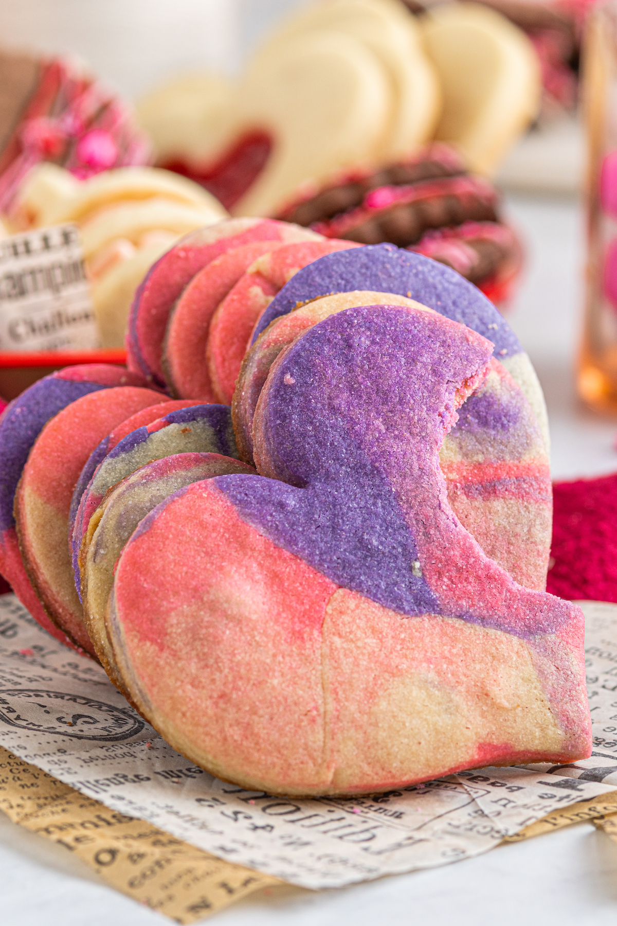 A stack of pink-and-purple heart-shaped cookies is leaning against a platter of assorted cookies. The first cookie in the stack has a bite taken out of it.