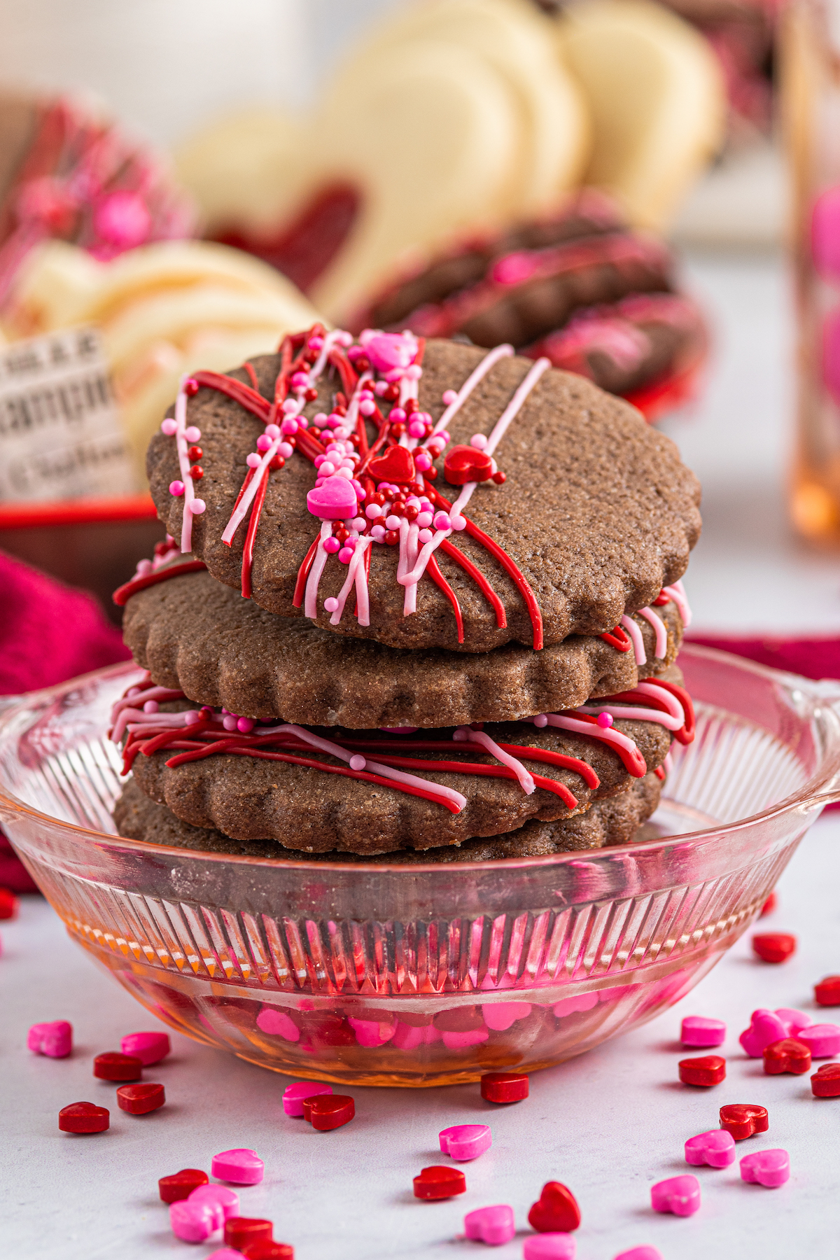 Four chocolate cut-out cookies stacked on top of each other, in a small cut-glass dish. Sprinkles are scattered around the dish on the table, and a platter of cookies is in the background of the shot.