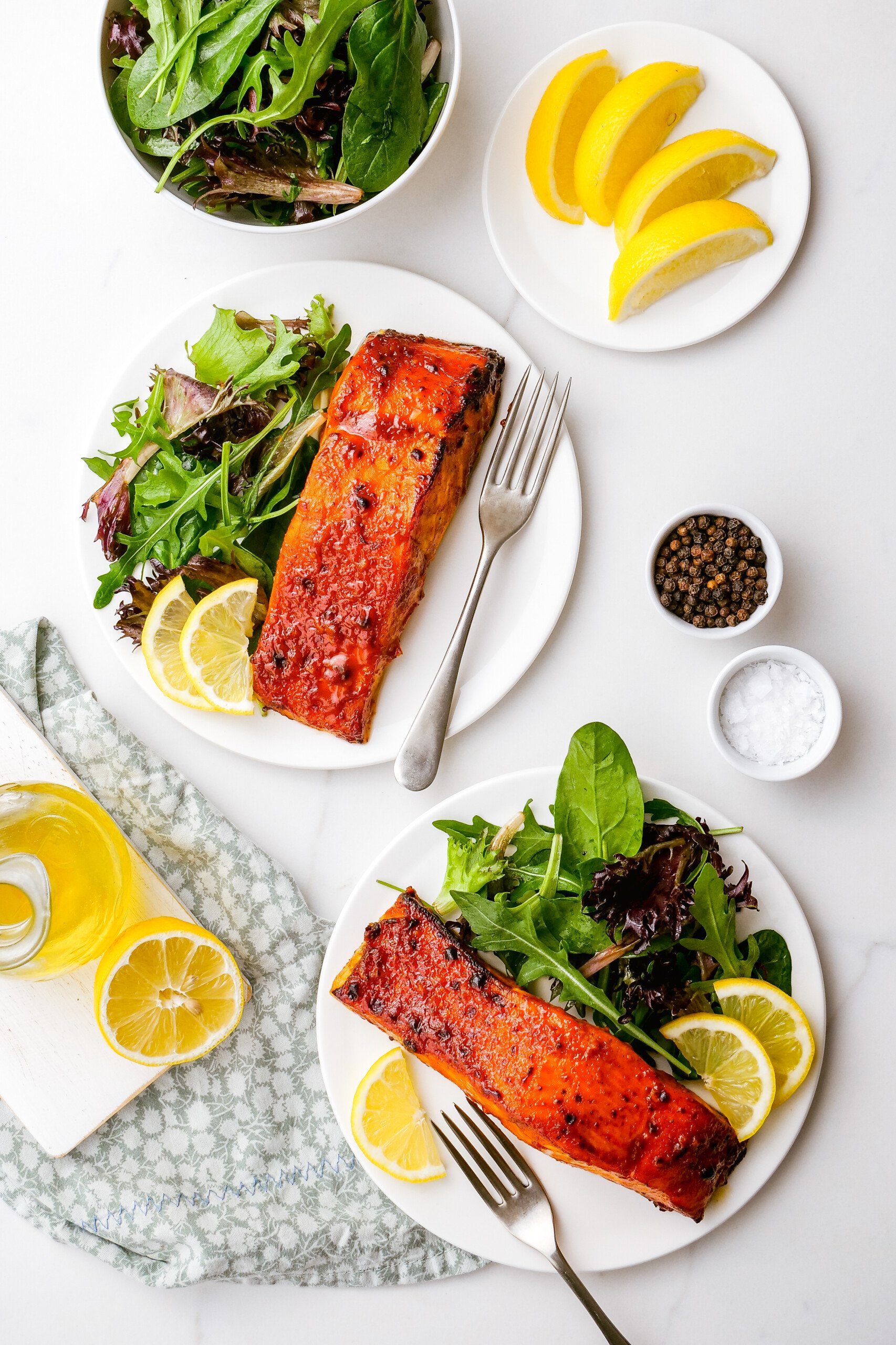 Overhead shot of a table with a small bowl of spring mix, a small dish of lemon wedges, and two dinner plates with servings of salmon, salad, and lemon garnish.