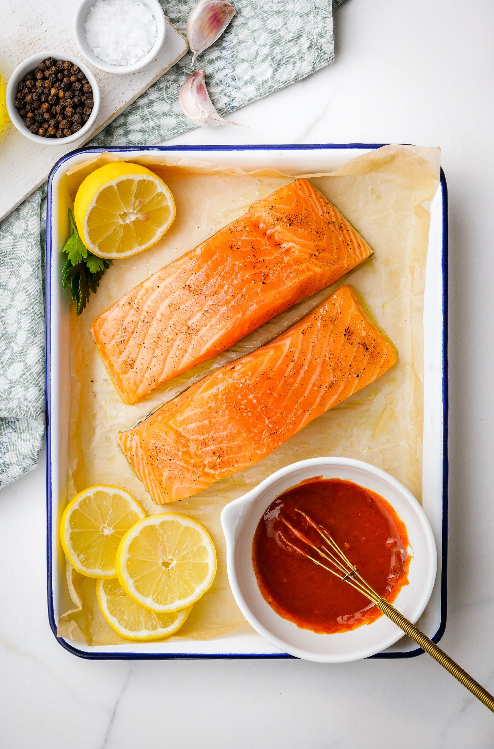 Uncooked salmon fillets o a parchment-lined baking sheet with lemon slices and a small bowl of sauce.