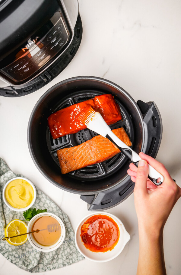 An air fryer with two fish fillets inside. A woman is brushing sauce over one of the fillets.