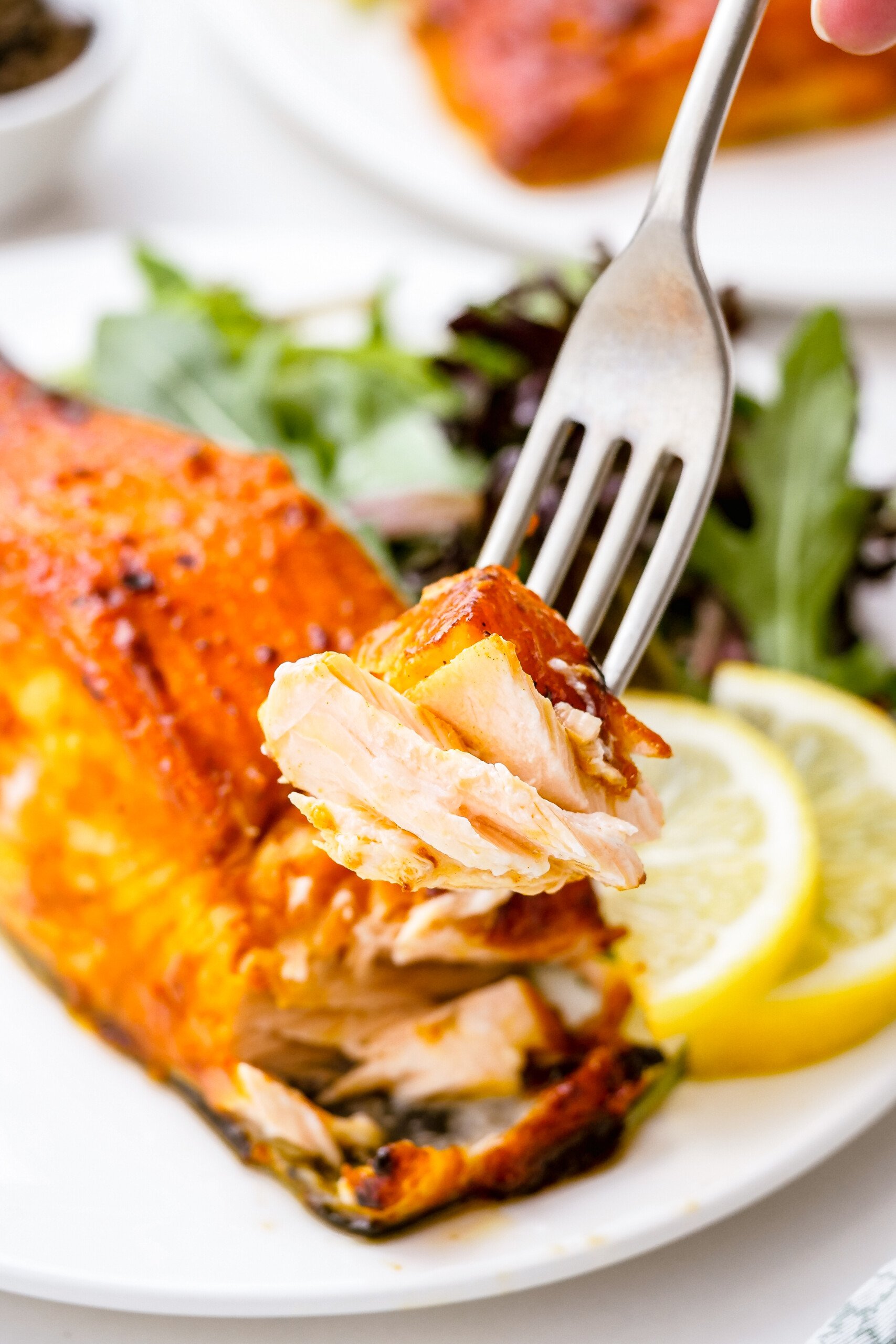 Close-up shot of a bite of fish on a fork. In the background is a dinner plate with the fish fillet and sides.