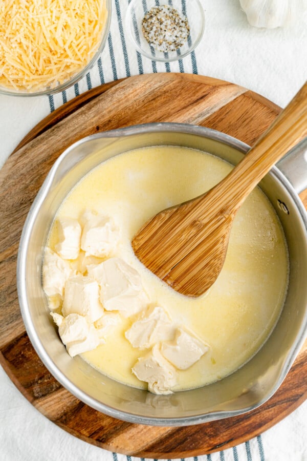A saucepan full of creamy sauce with cubes of cream cheese being stirred into it with a wooden spoon.