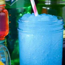 A blue vodka Slushies with a pink straw in a mason jar with jolly rancher candies around the bottom.