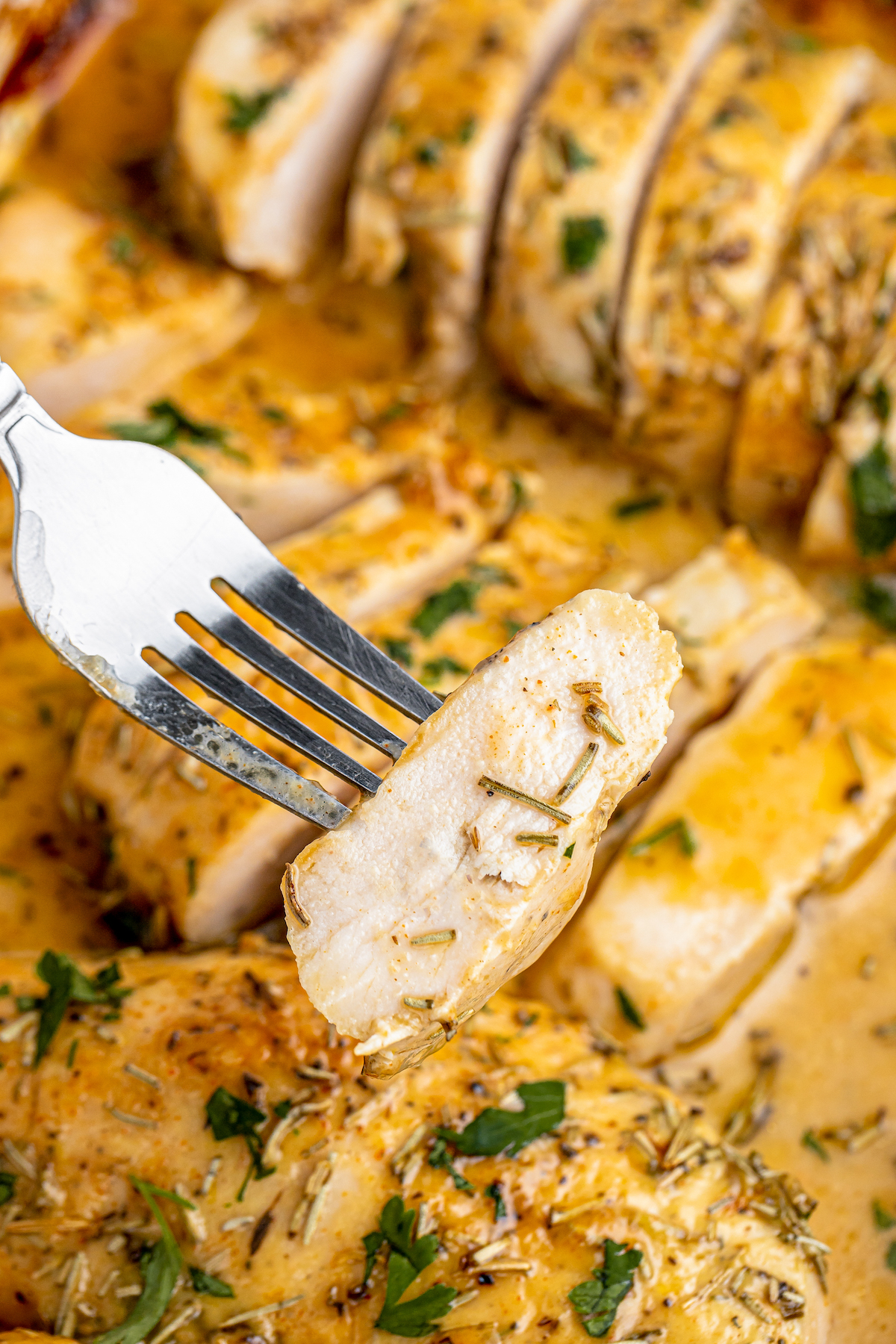 A fork lifting a piece of sliced chicken breast out of a baking dish of baked chicken.