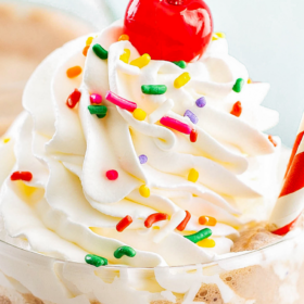 Up close image of a chocolate milkshake in a glass with whip cream, rainbow sprinkles and a red and white striped straw.