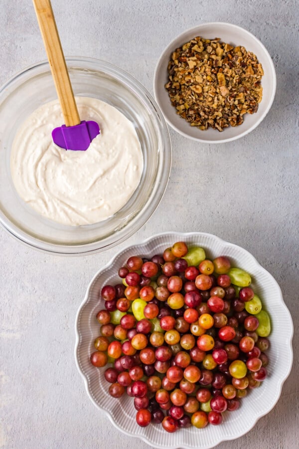 A bowl of grapes, a bowl of chopped nuts and toppings, and a bowl of creamy sauce with a spatula.