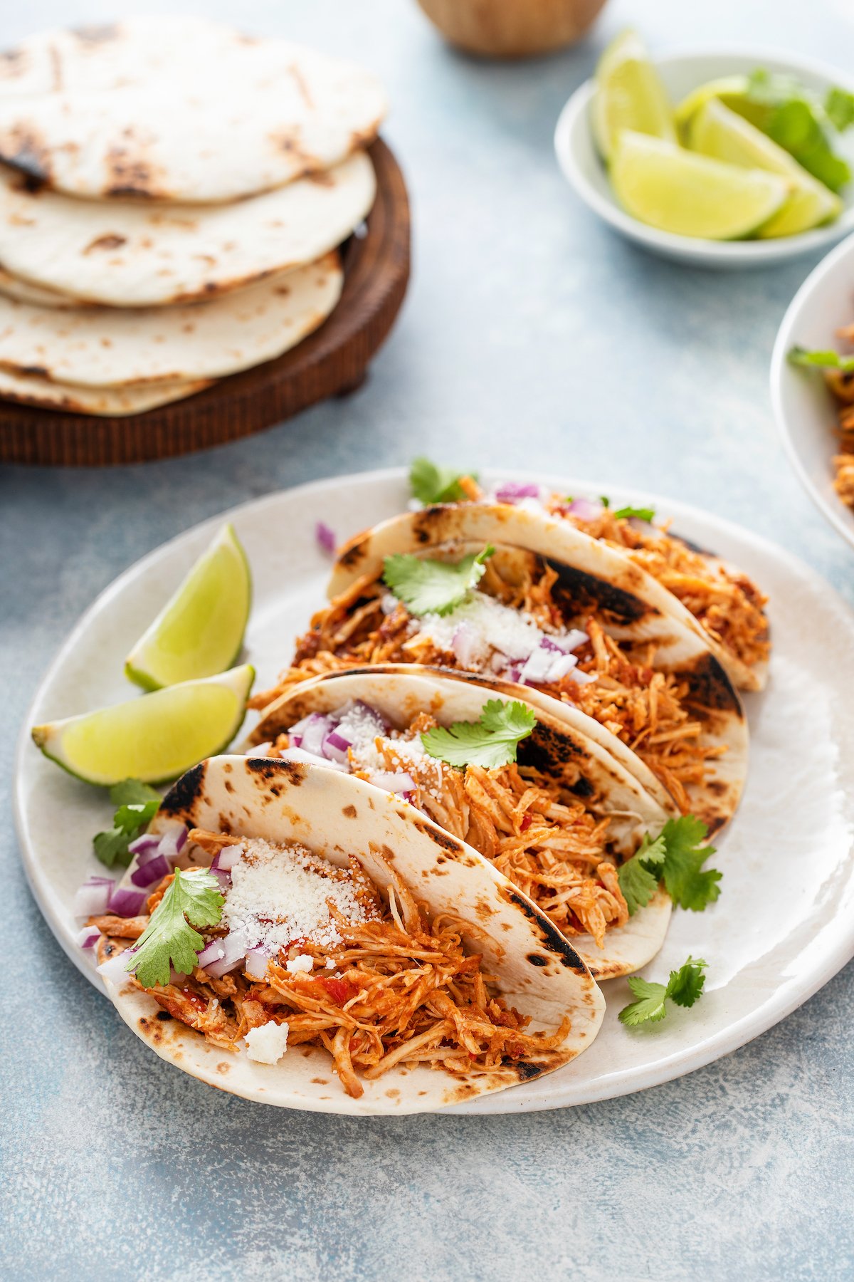 A plate of shredded chicken tacos next to a stack of tortillas and a dish of lime wedges.