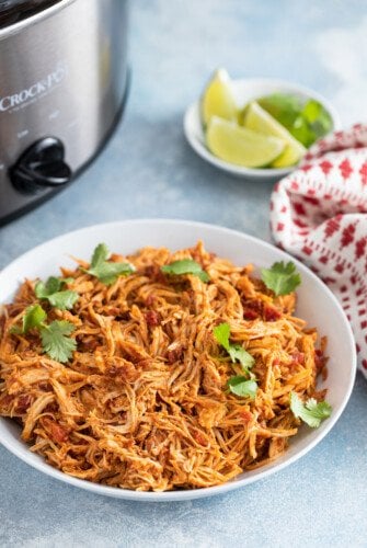 A bowl of shredded chicken next to a slow cooker and plate of lime wedges.