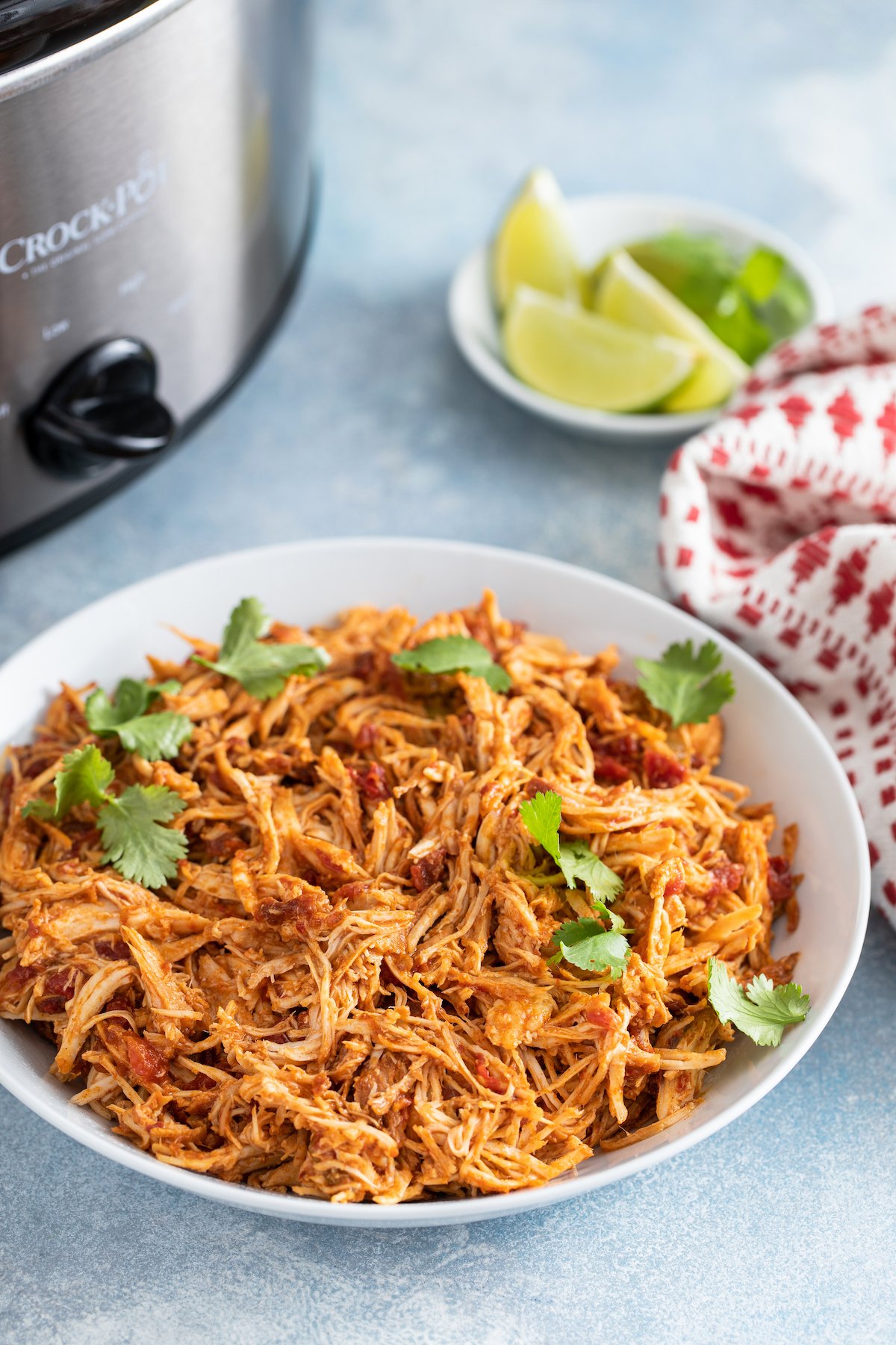 A bowl of shredded chicken next to a slow cooker and plate of lime wedges.
