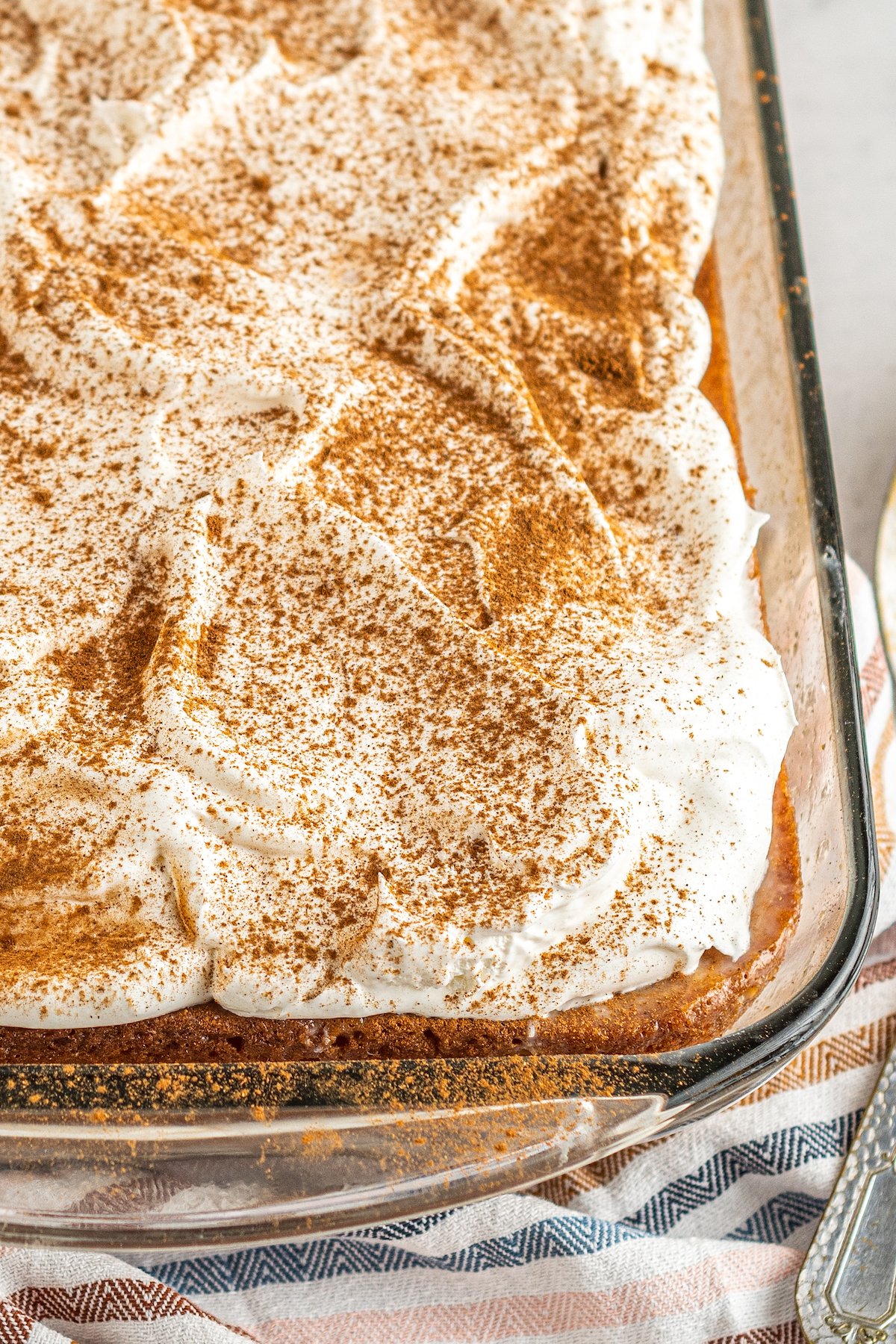 Close-up shot of the edge of a sheet cake in a glass pan, topped with whipped cream and cinnamon.