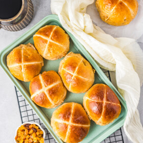 Six sweet Easter buns on a rectangular blue serving platter. Two more buns are on a white linen kitchen towel.