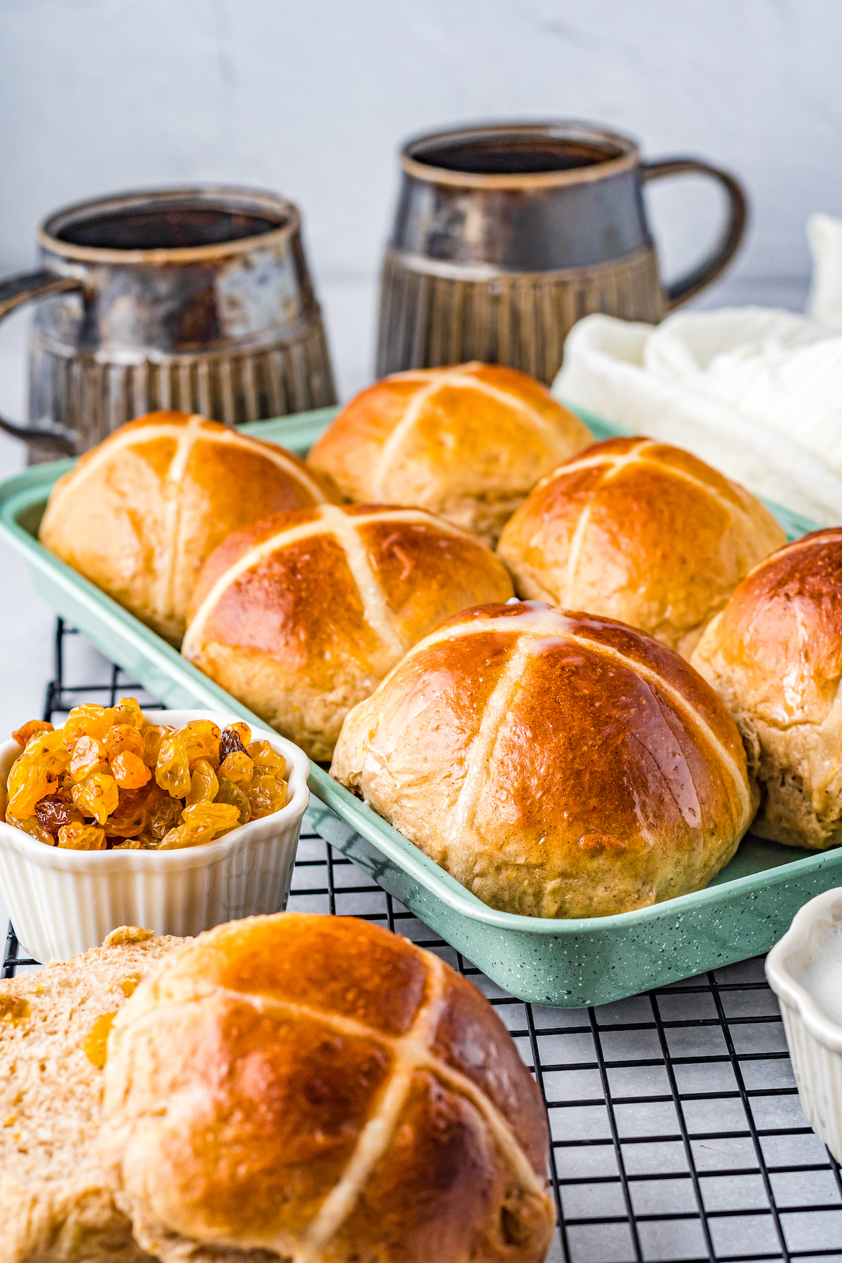 Six baked buns on a plate, cooling on a baking rack, with a bowl of golden raisins nearby.