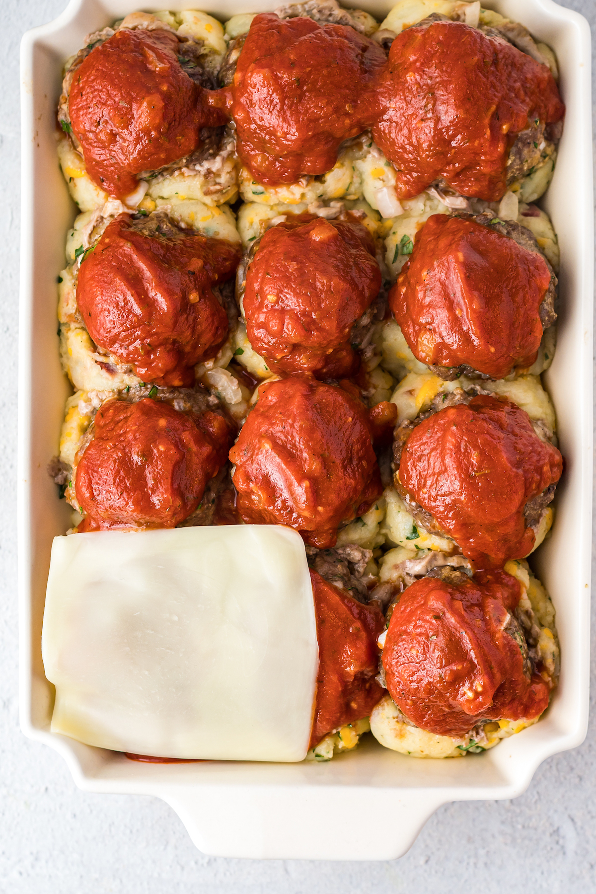 A baking dish with layers of mashed potato, meatballs, and sauce. A slice of cheese is laid on top of one corner of the dish.