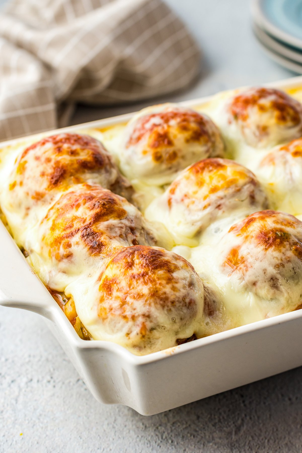 A rectangular white casserole dish topped with cheesy meatballs, marinara, and potatoes.