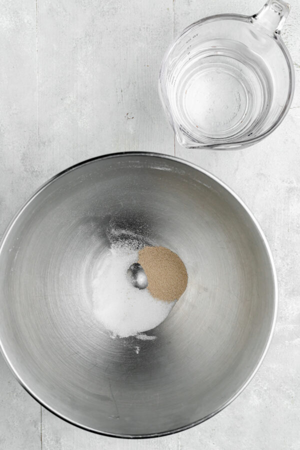 Yeast, sugar, and salt in a mixing bowl. A measuring cup of water is on the work surface as well.